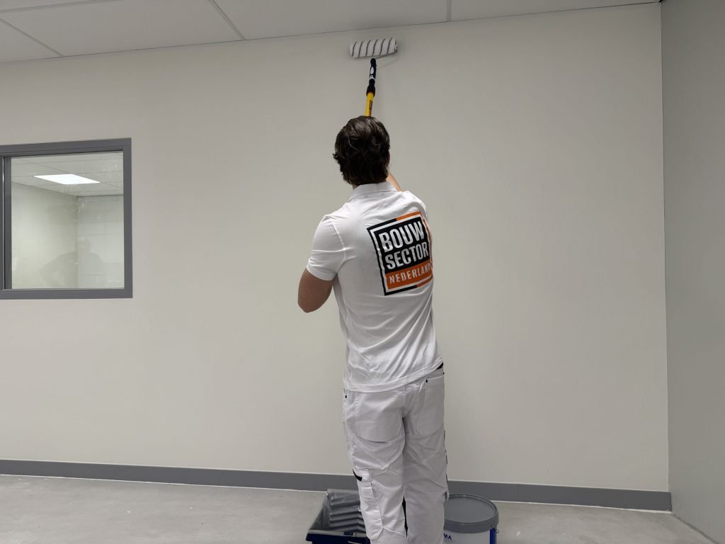 Schilder in witte werkkleding rolt een witte wand tot vlak onder het plafond, met de verfbak aan zijn voeten