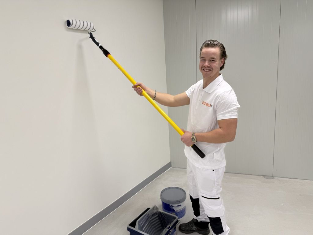 Lachende schilder met een roller aan een gele steel, naast een emmer Sigma-verf en een verfbak op de vloer