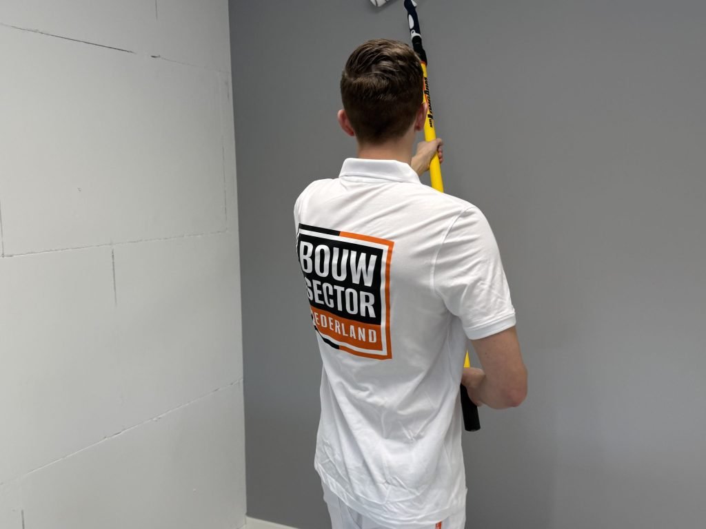 Schilder met Bouwsector-logo op de rug werkt een donkergrijze wand af naast een ongepleisterde blokkenwand