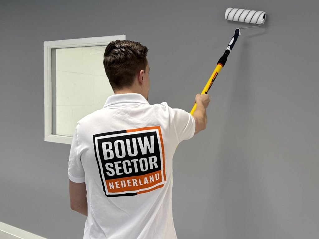 Schilder van Bouwsector Nederland zet met een verlengde roller grijze verf op een wand naast een raam