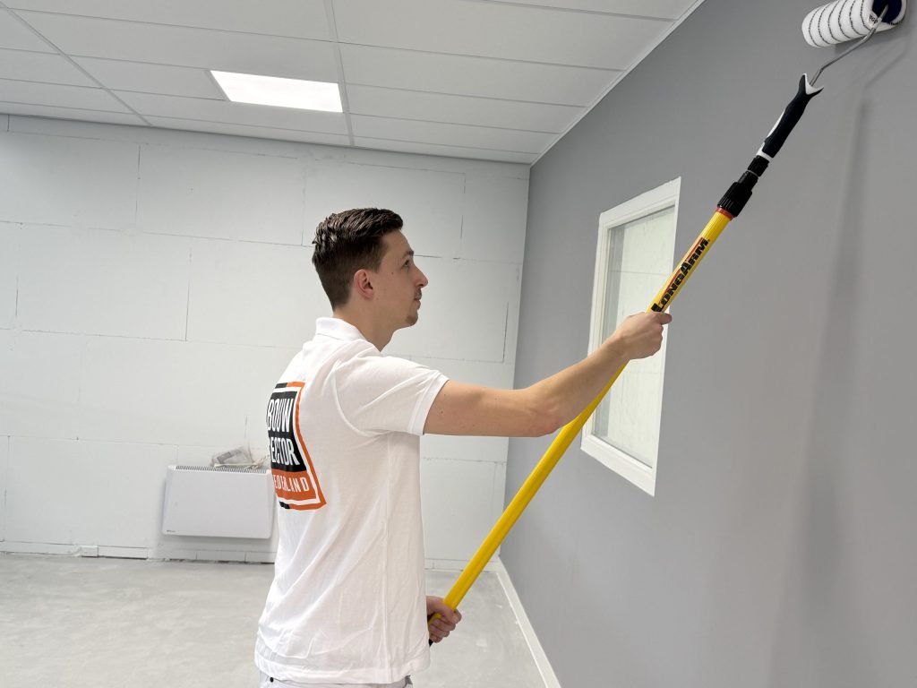Schilder in wit shirt zet de roller hoog op een grijze wand bij een raam, achter hem een kale muur van blokken