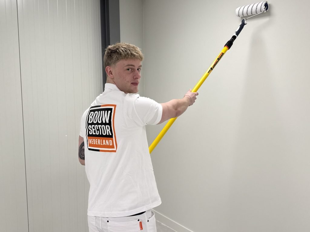 Schilder van Bouwsector Nederland kijkt om terwijl hij met een lange roller een lichte muur afwerkt