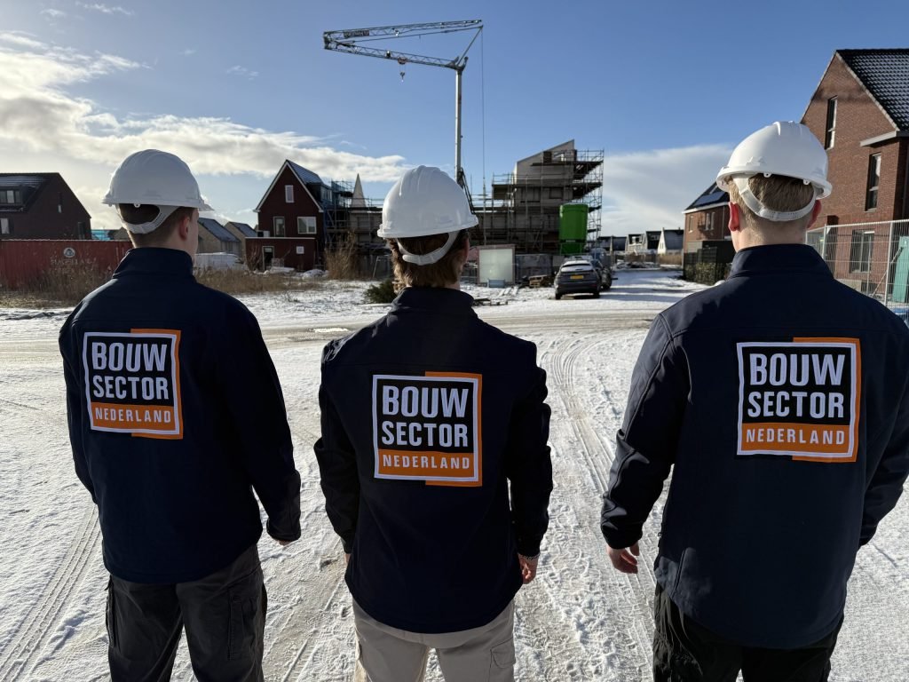 Drie vakmensen van Bouwsector Nederland met witte helm op een besneeuwde straat, met een kraan en nieuwbouw in aanbouw