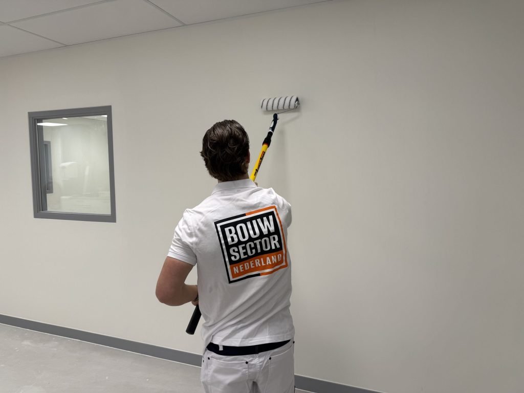 Schilder van Bouwsector Nederland rolt met een verlengsteel een lichte wand naast een binnenraam